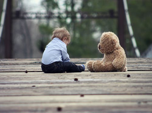Free Adorable toddler sitting with a teddy bear on a wooden bridge, enjoying a peaceful moment outdoors. Stock Photo