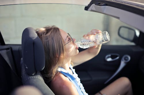 Free High angle side view of young woman in glasses and casual clothes drinking water from plastic transparent bottle while sitting in cabriolet with open roof in traffic jam in hot sunny day Stock Photo