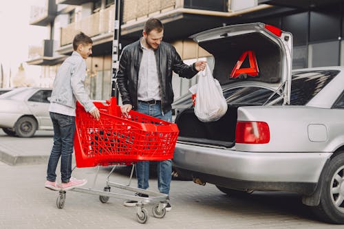 Free Full body of happy man and boy in casual clothes moving shopping bag from plastic trolley to car trunk on parking near modern buildings Stock Photo